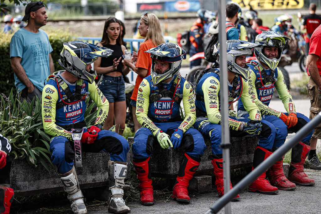 Four Sherco Team EnduroGP riders sat on a wall in the foreground in their blue and green sponsored clothing