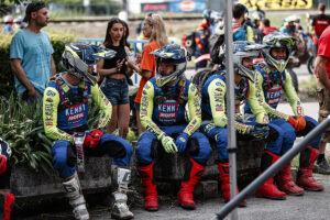 Four Sherco Team EnduroGP riders sat on a wall in the foreground in their blue and green sponsored clothing